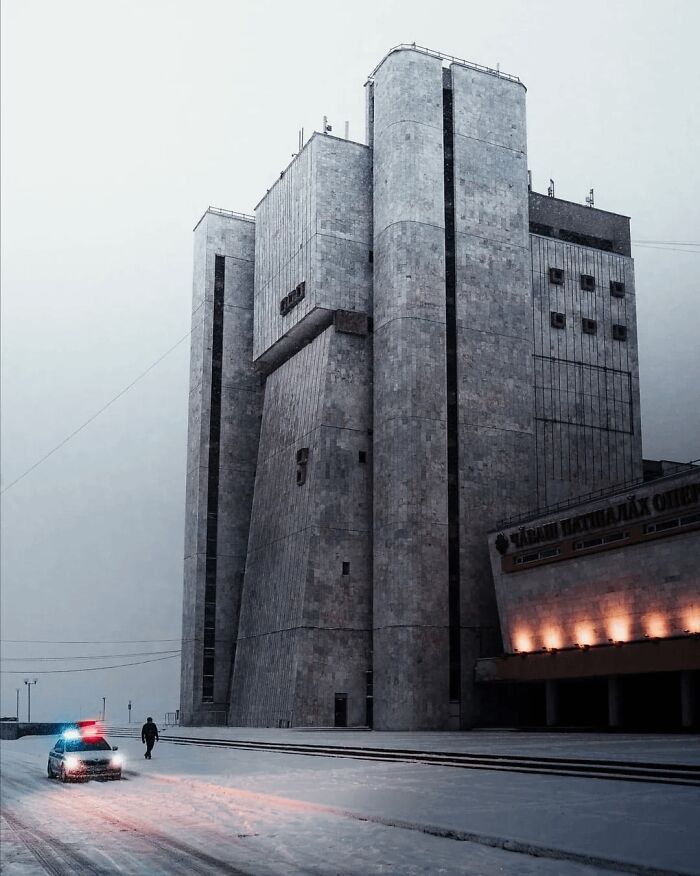 Massive brutalist building with a police car and lone figure in snowy, dystopian nightmare-like urban setting at dusk.
