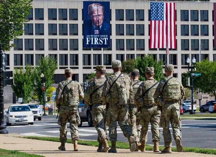 National guardsmen walking past a building with a large political banner and American flag in a dystopian nightmare setting.