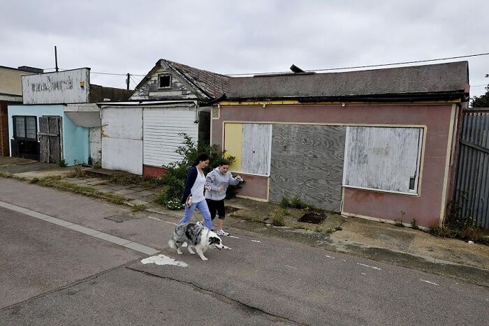 Two people walking a dog past boarded-up buildings in a scene resembling a dystopian nightmare.