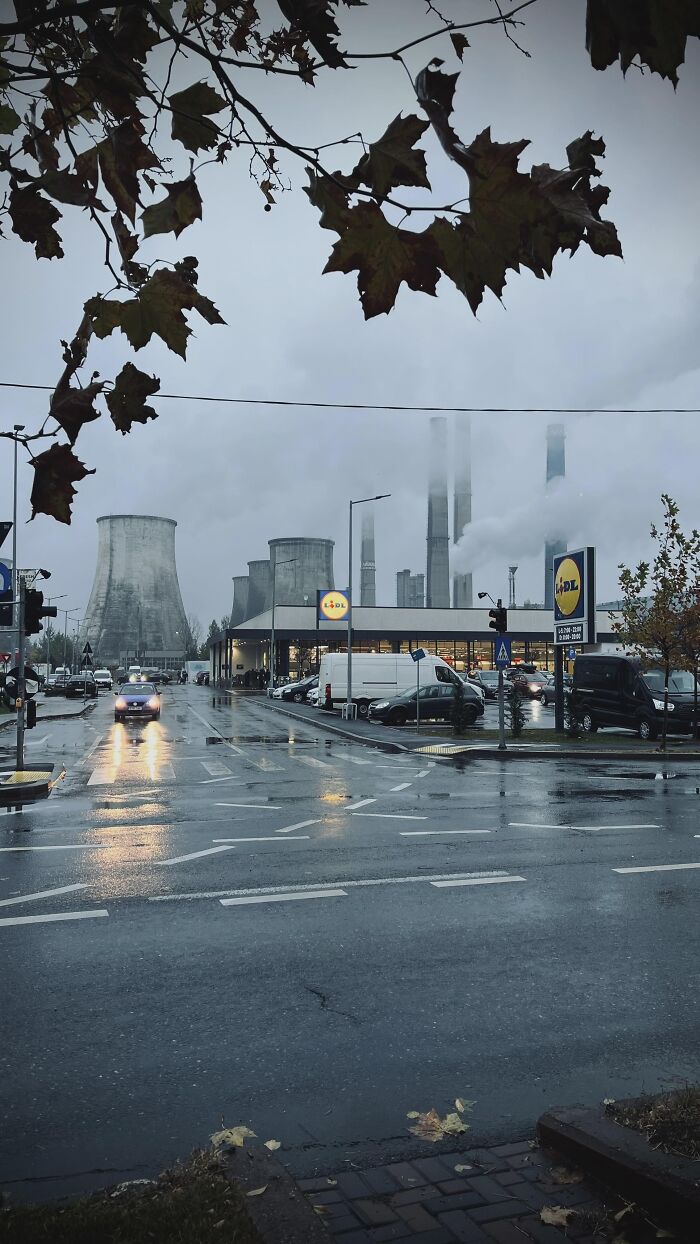 Industrial complex emitting smoke behind a supermarket on a rainy day, evoking dystopian nightmare atmosphere.