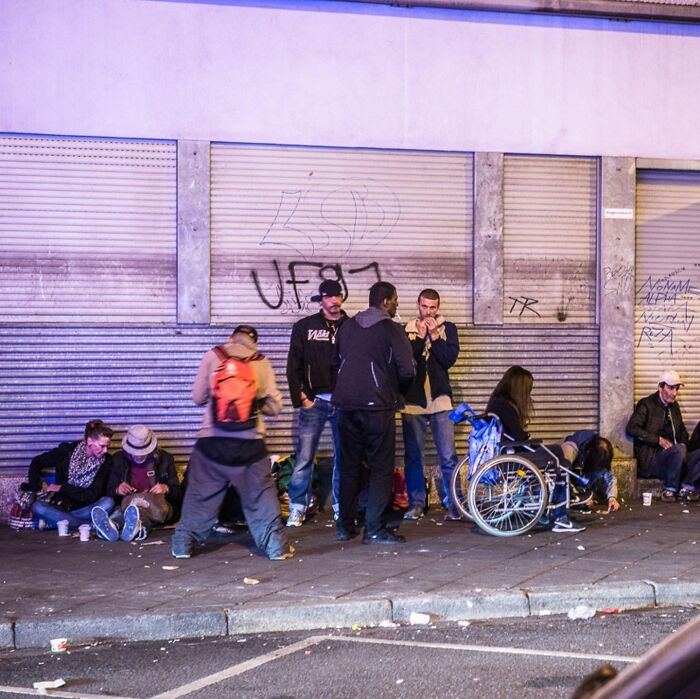 Group of people, including a person in a wheelchair, gathered on a dirty urban street with graffiti, evoking a dystopian nightmare scene.