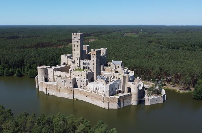 A large, fortress-like castle surrounded by water and dense forest, evoking a dystopian nightmare atmosphere.