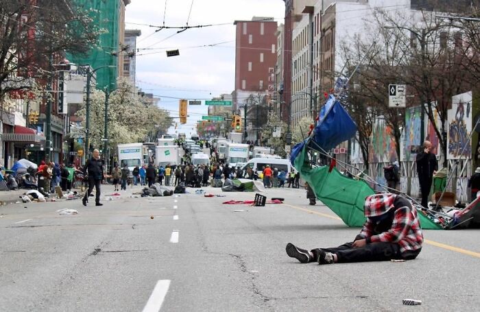 Person sitting on an empty street amid debris and tents in a chaotic urban scene resembling a dystopian nightmare.