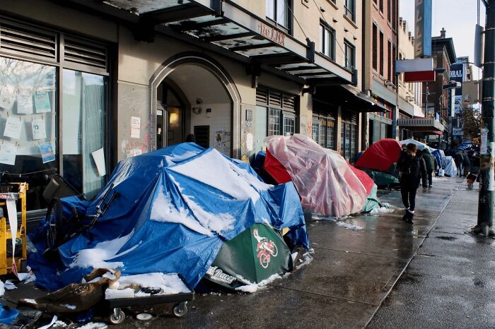 Tents covered in snow lined up on a city sidewalk, depicting a dystopian nightmare of urban homelessness.