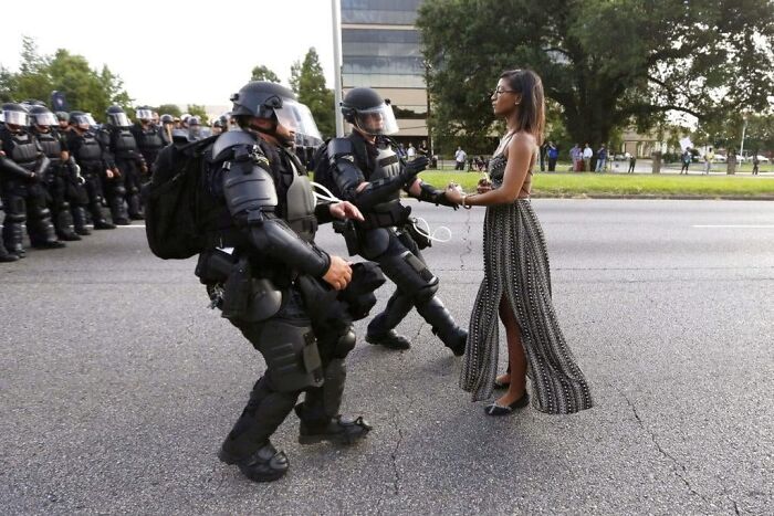 Woman calmly facing riot police in full gear on a city street, a dystopian nightmare scene from around the world.