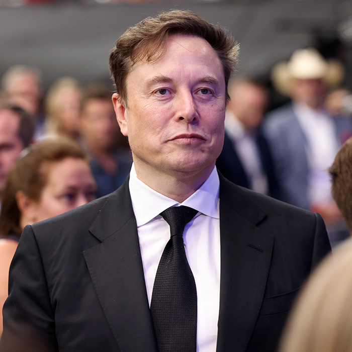 Elon Musk in a black suit at an event, with people blurred in the background discussing UFO files and Trump.