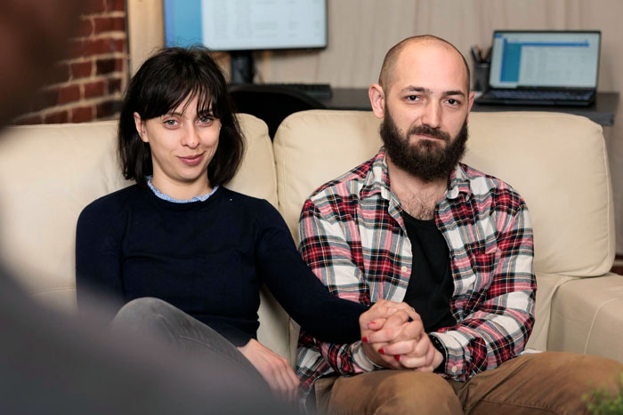 A couple sitting closely on a beige sofa holding hands, reflecting an embarrassing step mother relationship affair moment.
