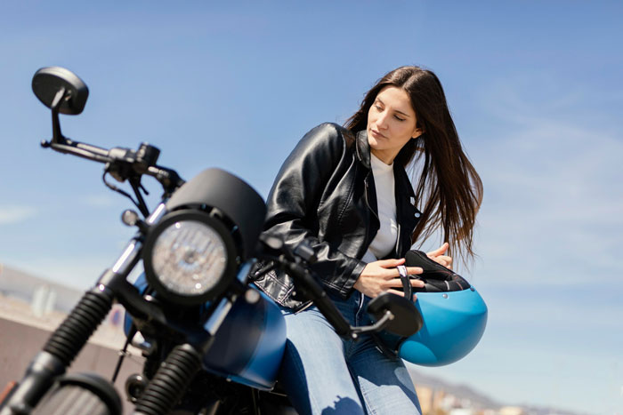 Woman in a leather jacket holding a blue helmet next to a motorcycle, related to selling bike spare parts profit.