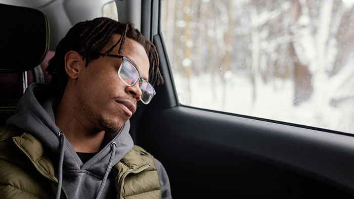 Young man with glasses and hoodie resting in a car seat looking out the window, reflecting on family and respect issues. Young man with glasses and hoodie resting in a car seat looking out the window, reflecting on family and respect issues.