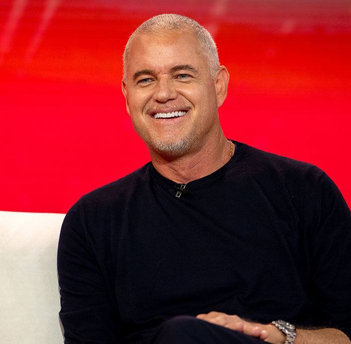 Eric Dane smiling during an interview, wearing a black shirt against a bright red background.