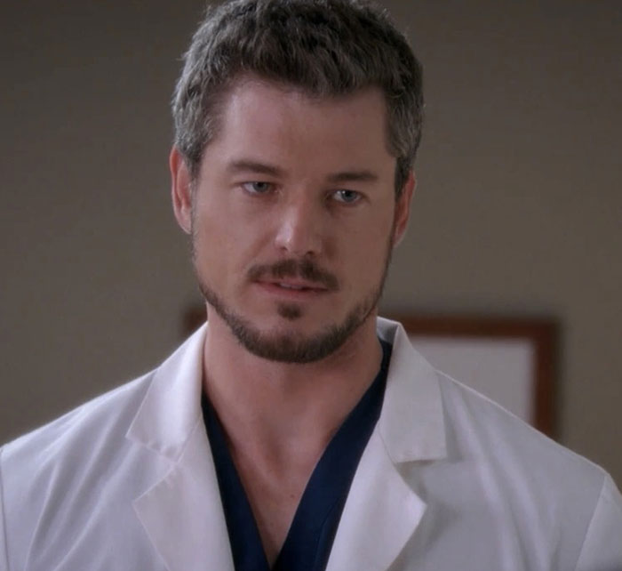 Eric Dane in a Grey's Anatomy scene, wearing a white coat and dark scrubs, with a serious expression on set.