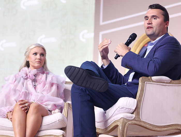 Man in blue suit speaking into microphone while seated next to woman in pink dress amid Epstein link conspiracy discussion.