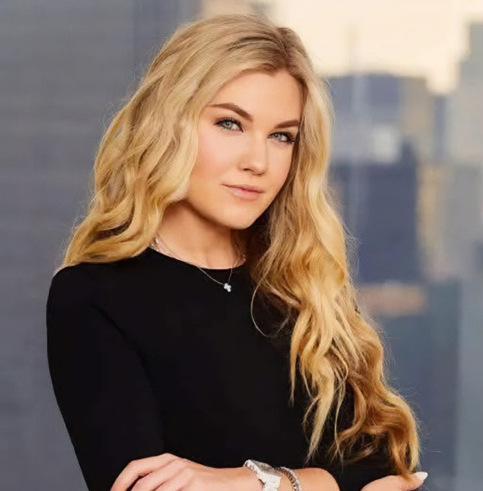 Woman with long blonde hair wearing a black top, posing confidently against a blurred cityscape background, Epstein link discussed