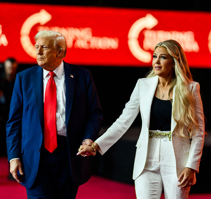 Erika Kirk in a white suit holding hands with a man in a blue suit and red tie at a public event.