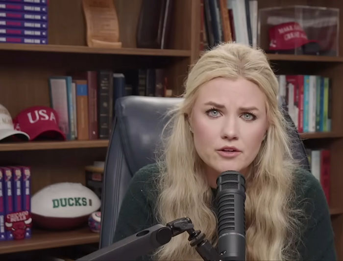 Blonde woman speaking into a microphone in an office with shelves, books, and sports memorabilia behind her, Erika Kirk.