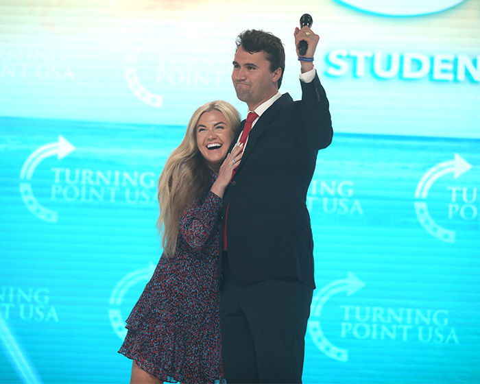 Erika Kirk and Charlie Kirk on stage at Turning Point USA event, with bright blue background and microphone in hand.