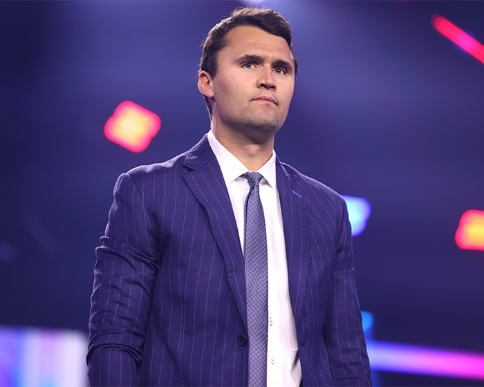 Man in a dark pinstripe suit standing on stage with colorful blurred lights in the background at an event.