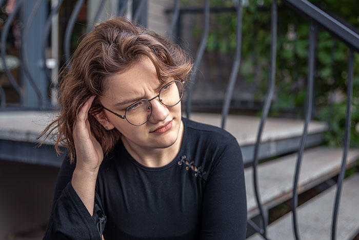 Young woman wearing glasses looks concerned while sitting outdoors near metal stairs, relating to K-Pop fan rare merch conflict.