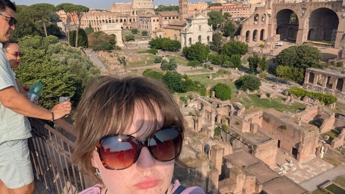 Woman taking a chaotic mom selfie with large sunglasses overlooking ancient ruins and historic architecture in the background.