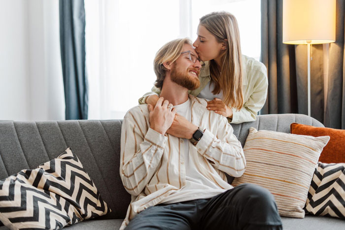 Woman kisses her high school bestie on the forehead as they share a cozy moment on a couch, highlighting their growing love.