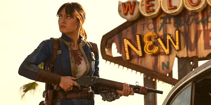 Woman in a blue jumpsuit holding a rifle standing near a worn Welcome to New Vegas sign in a post-apocalyptic setting Fallout Season 3 - 1