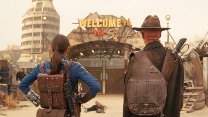 Two characters with backpacks face the Welcome to the Strip sign in a desert setting from Fallout Season 3. - 4