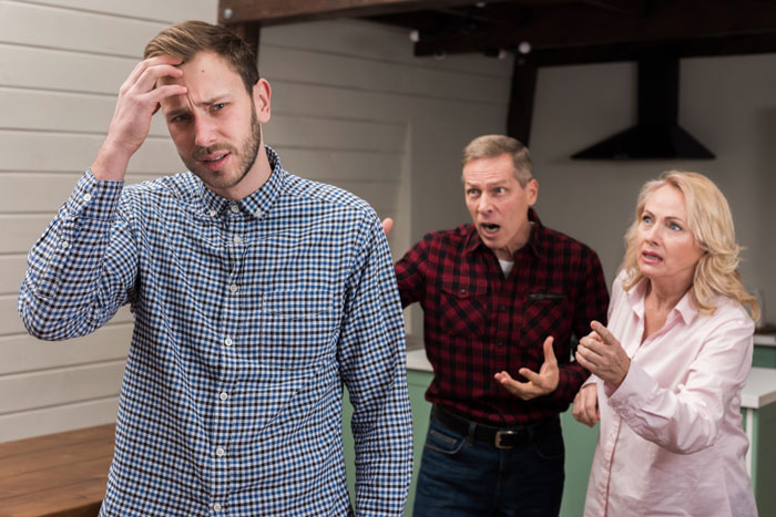 Young man stressed while parents argue in the background, highlighting effects of parents' favoritism on children behavior.