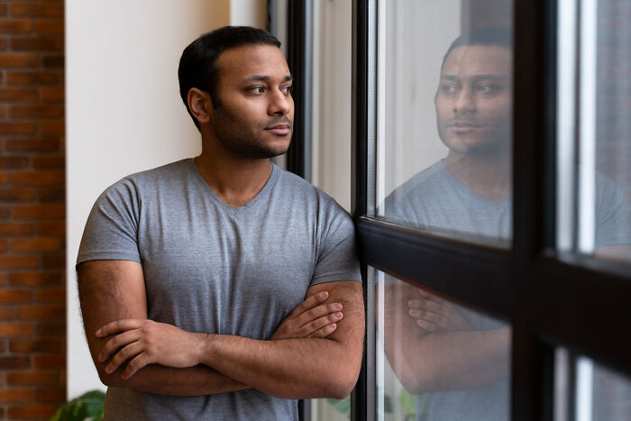 Young man in gray shirt looking pensively out a window, reflecting on the emotional reality of being related to an ICE agent.