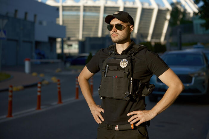 ICE agent standing confidently outdoors wearing tactical gear and sunglasses, representing the emotional reality of related families.