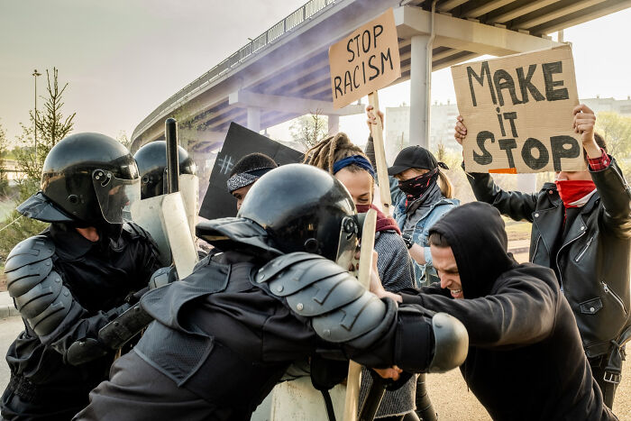 Protesters confront riot police under a highway, holding signs against racism and injustice related to ICE agents.