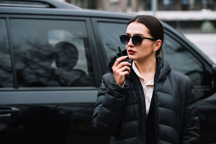 Woman wearing sunglasses and black jacket speaking on a radio near a black vehicle, symbolizing emotional reality of ICE agent family.