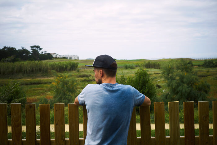 Man in a blue shirt and cap looking over a fence at a green field, reflecting the emotional reality of being related to an ICE agent