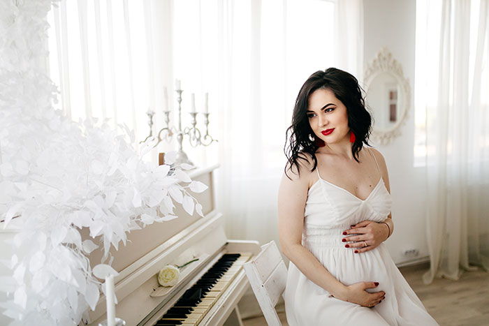 Pregnant woman in white dress sitting by piano, reflecting on family patriarch and brothers&rsquo; marriages collapsing.