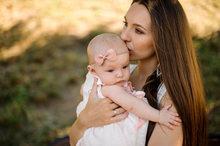 Woman with long brown hair holding and kissing a baby girl with a pink bow, reflecting baby fever and pregnancy emotions.