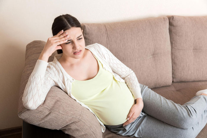 Pregnant woman with baby fever sitting on a couch, looking tired and overwhelmed with her pregnancy symptoms.