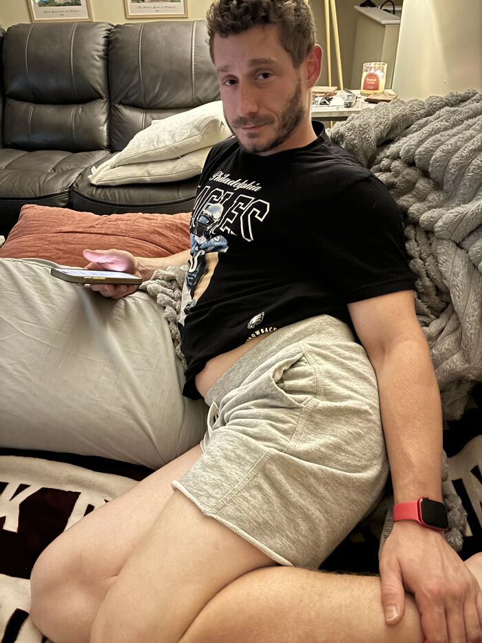 Man lounging awkwardly on couch holding phone, wearing casual shorts and a black graphic t-shirt, showing uncomfortable body language