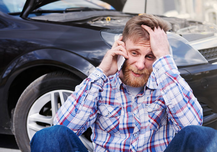 Man sitting by broken car with hood open, stressed and on phone, reacting to sudden $500 insurance hike and fianc&eacute;e's frustration.