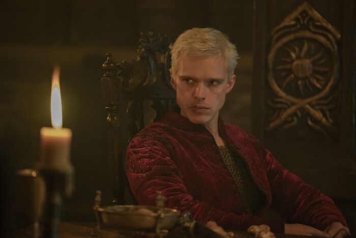 A Knight Of The Seven Kingdoms villain sitting in a dark room wearing a red outfit near a lit candle. A Knight Of The Seven Kingdoms villain sitting in a dark room wearing a red outfit near a lit candle.