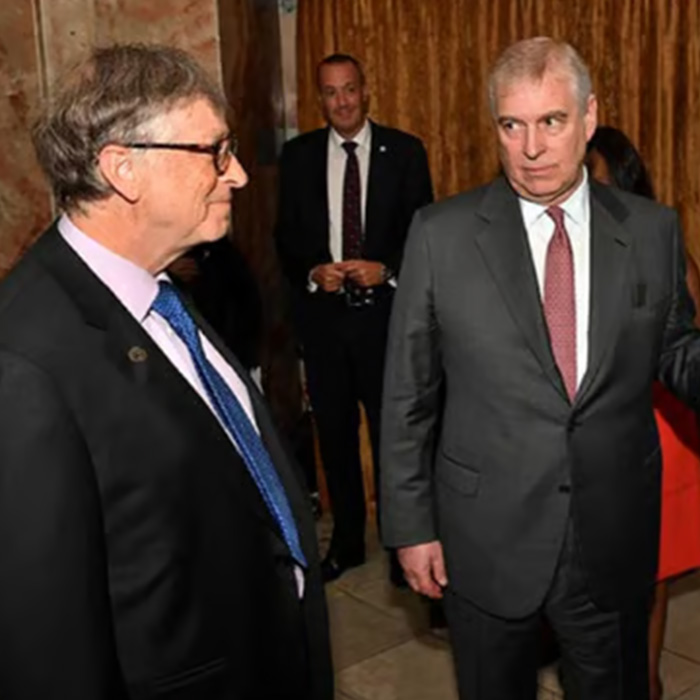 Prince Andrew wearing a suit with a terrified look, standing indoors near two other men in formal attire.