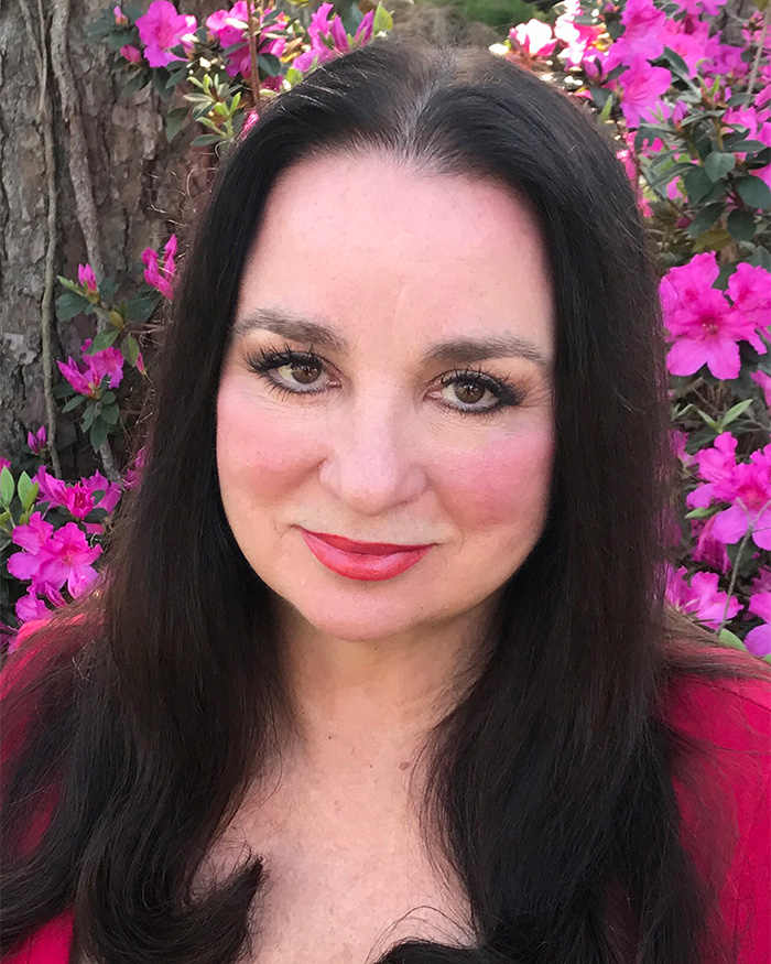 Nancy Guthrie smiling outdoors with dark hair and pink flowers in the background in a natural setting.
