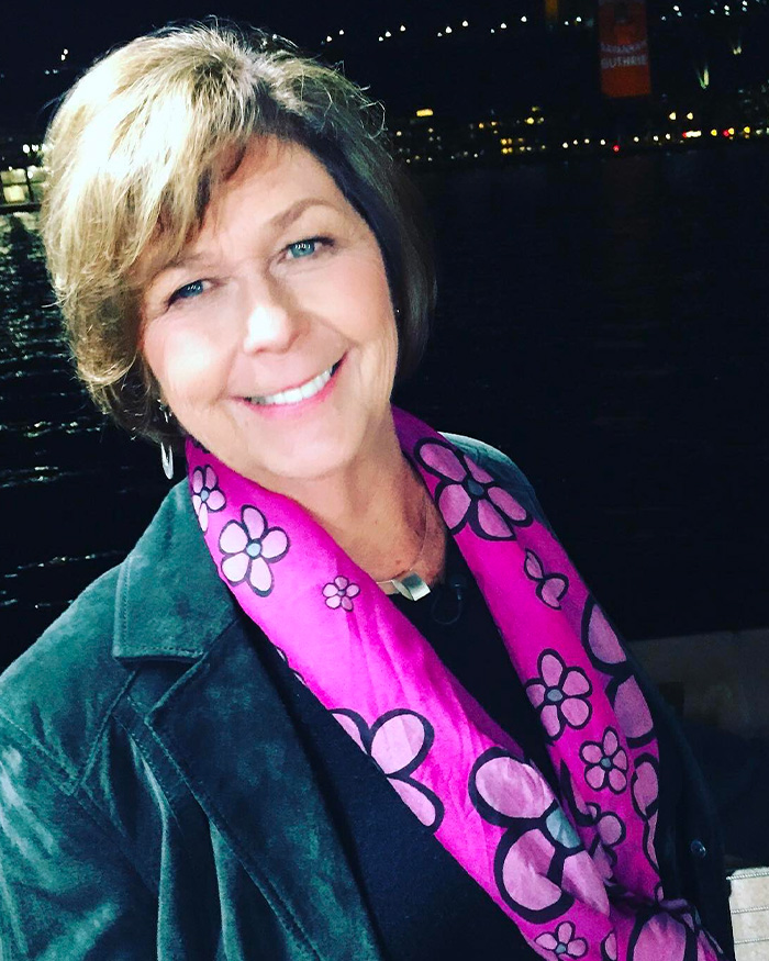 Woman smiling outdoors at night, wearing a pink floral scarf and dark jacket, related to Nancy Guthrie masked suspect sketch.