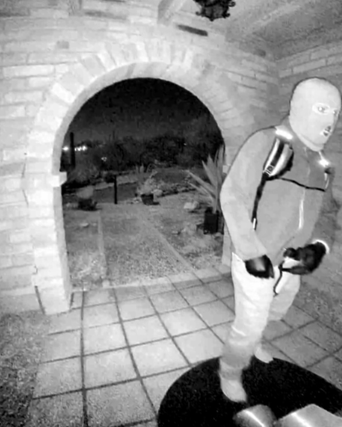 Masked suspect captured on security camera at night near brick archway and tiled pathway in a residential area.