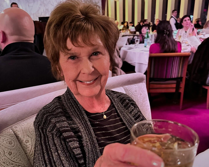 Elderly woman smiling and raising a glass in a restaurant setting with people dining in the background.
