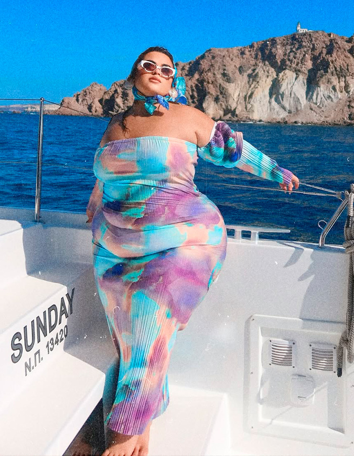 Former body positivity influencer posing confidently on a boat wearing a colorful dress with ocean and cliffs in background