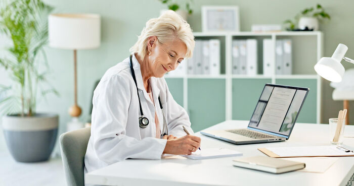 Female physician writing notes at desk with laptop in bright office, illustrating hilarious physicians notes for daily laughter.