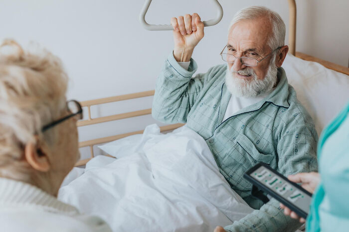 Elderly male patient in hospital bed using support handle, smiling at visitors during a lighthearted physicians' notes moment.