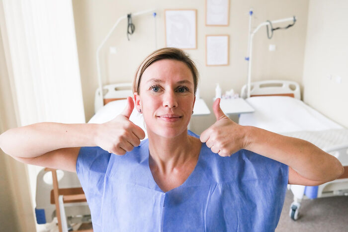 Woman in hospital gown giving thumbs up in a patient room, related to hilarious physicians' notes for daily laughter.
