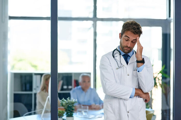Male physician in white coat looking stressed indoors beside glass wall with colleagues in background featuring hilarious physicians notes theme.