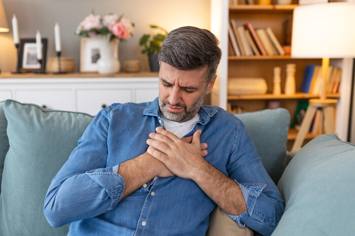 Man in a blue shirt sitting on a couch, holding his chest, illustrating a scene related to hilarious physicians' notes.