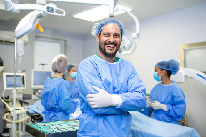 Smiling physician in surgical scrubs with team in background preparing for operation in a bright medical room.